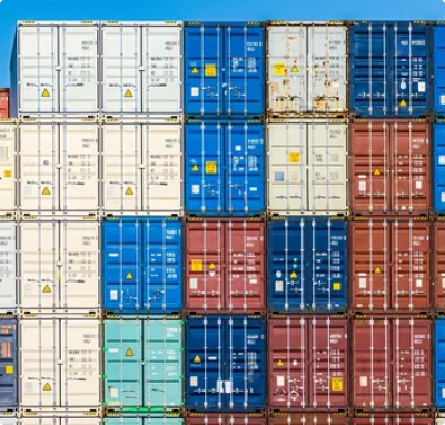 Stacked shipping containers in multiple colors at a port or industrial logistics yard, representing heavy industry and global trade.