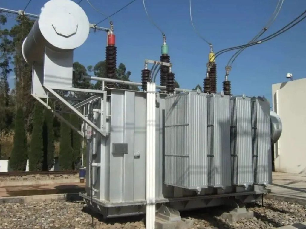 High-voltage power transformer installed at an electrical substation with bushings, cooling radiators, and overhead transmission connections.